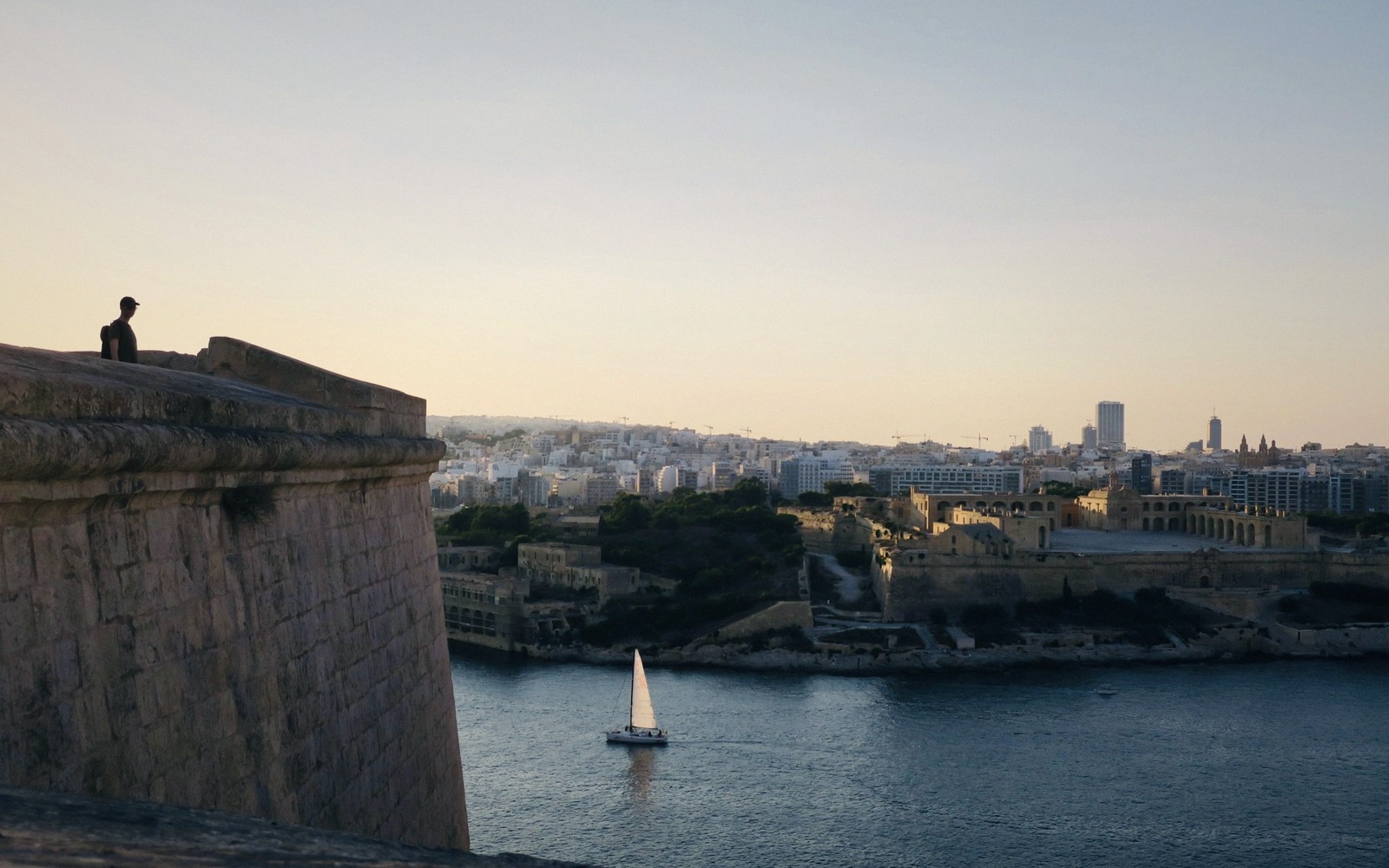 A person sits atop a stone bastion overlooking a harbor with a sailboat and coastal city