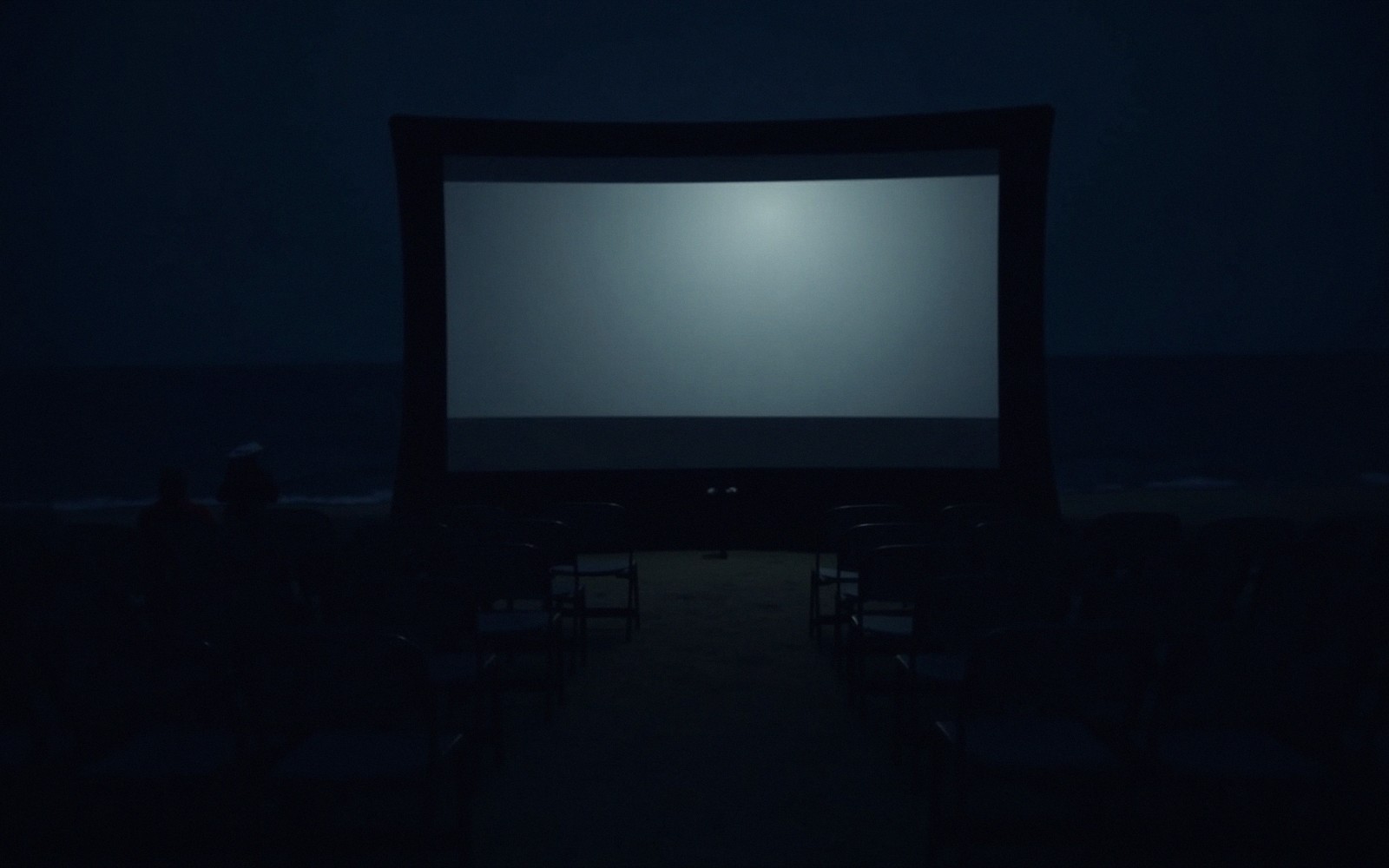 Rows of empty chairs face a glowing inflatable screen on a dark beach at night